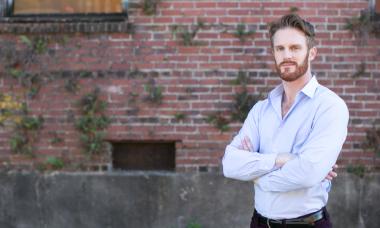 A white man with brownish ginger hair and beard wearing a button up shirt stands in front of a brick wall with vines climbing. His arms are crossed and his expression is soft.