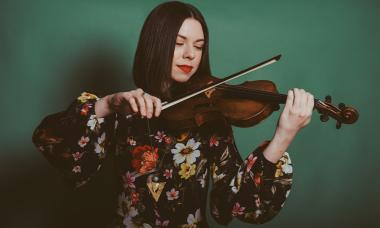 A white brunette feminine person with a blunt bob cut, red lipstick, wearing a floral long sleeve shirt, plays the violin.