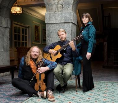 A woman, standing; a blonde man with a guitar, seated; a bearded man with red hair and a violin seated on the floor.