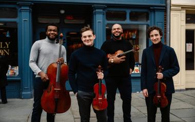 The Isidore String Quartet stands in front of a blue building, each holding their instruments