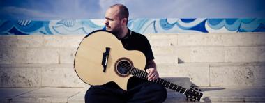 Andy is seated on marble steps, holding a bblond acoustic guitar on his lap, and looks to the side.