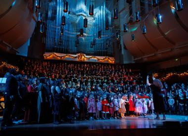 A choir of girls, young women, and non-binary singers hundreds strong perform onstage at a festively decorated Davies Symphony Hall, conducted by Valérie Sainte-Agathe, in low blue lighting.