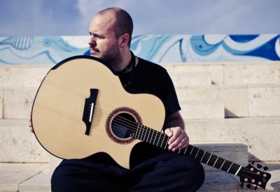 Andy is seated on marble steps, holding a bblond acoustic guitar on his lap, and looks to the side.